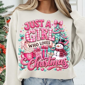 May include: A cream-colored sweatshirt featuring a graphic print with the text "JUST A GIRL WHO LOVES CHRISTMAS". The design includes a pink plaid font, a decorated Christmas tree, a snowman with a plaid scarf, gift boxes, and a mug of cocoa.