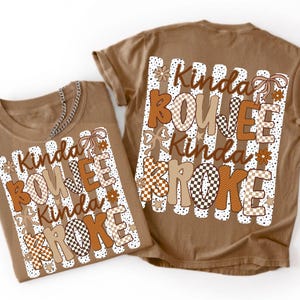 Pode incluir: Uma t-shirt castanha clara com uma impress&atilde;o gr&aacute;fica com o texto Kinda BOUJEE Kinda BROKE em padr&otilde;es castanhos e xadrez. O design inclui riscas verticais brancas, pequenos pontos, flores castanhas e ilustra&ccedil;&otilde;es de la&ccedil;os.