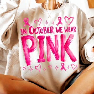 May include: A beige crewneck sweatshirt featuring the pink text "IN OCTOBER WE WEAR PINK" in a brushstroke font. The design includes pink ribbon symbols, heart outlines, and small four-pointed star accents scattered above and below the text.
