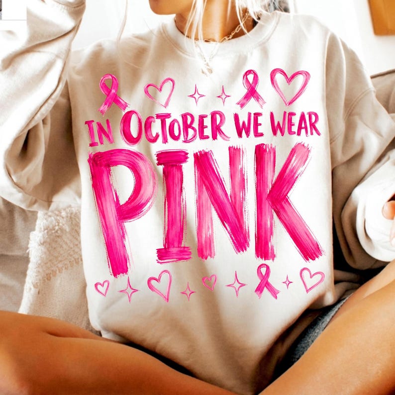 May include: A beige crewneck sweatshirt featuring the pink text "IN OCTOBER WE WEAR PINK" in a brushstroke font. The design includes pink ribbon symbols, heart outlines, and small four-pointed star accents scattered above and below the text.