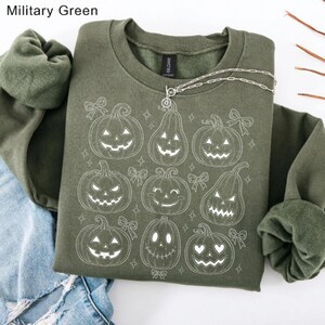 May include: A military green crewneck sweatshirt featuring a white line art graphic of nine various jack-o'-lantern pumpkins and small bows. The design includes pumpkins with different facial expressions, including triangle eyes and heart-shaped eyes.