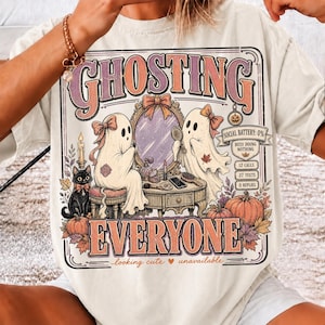 May include: An off-white t-shirt featuring a graphic of two ghosts at a vanity with a black cat, pumpkins, and autumn leaves. The text reads GHOSTING EVERYONE, SOCIAL BATTERY: 0%, BUSY DOING NOTHING, 12 CALLS, 27 TEXTS, 0 REPLIES, and looking cute unavailable.