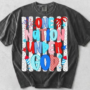 Pu&ograve; includere: Una t-shirt grigio antracite a maniche corte con una stampa grafica recante il testo ONE NATION UNDER GOD. Il design include motivi a scacchi e a pois rossi, bianchi e blu, illustrazioni di fuochi d'artificio e un fiocco rosso su sfondo a righe.