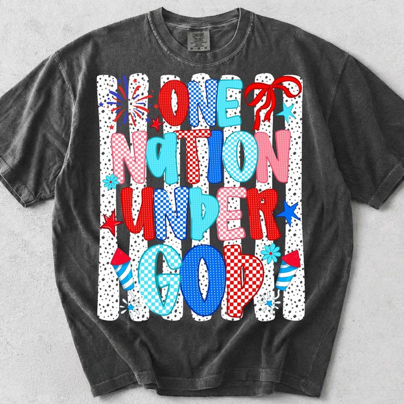 Pu&ograve; includere: Una t-shirt grigio antracite a maniche corte con una stampa grafica recante il testo ONE NATION UNDER GOD. Il design include motivi a scacchi e a pois rossi, bianchi e blu, illustrazioni di fuochi d'artificio e un fiocco rosso su sfondo a righe.
