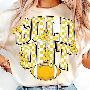 Puede incluir: Una camiseta color crema con el texto GOLD OUT en letras grandes con un patr&oacute;n de cuadros amarillos y blancos. El dise&ntilde;o incluye cintas de concienciaci&oacute;n amarillas integradas en las letras y un gr&aacute;fico de un bal&oacute;n de f&uacute;tbol americano abajo.