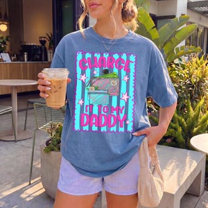 May include: A blue short-sleeve t-shirt featuring a graphic with vertical teal stripes, pink stars, and the text "CHARGE IT TO MY DADDY" in pink. The graphic includes illustrations of a wallet and credit cards with small white flower accents.