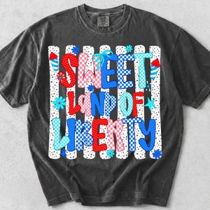 K&ouml;nnte beinhalten: Ein anthrazitfarbenes Kurzarm-T-Shirt mit einem Grafikdruck, der den Text SWEET LAND OF LIBERTY in rot-wei&szlig;-blau karierten Mustern zeigt. Das Design umfasst vertikale Streifen, Punkte, Feuerwerksk&ouml;rper und kleine Blumensymbole.