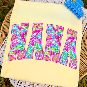 以下が含まれることがあります： 明るい黄色のTシャツに、大きなカラフルな文字で「NANA」と書かれています。文字はピンク、青、緑の色合いの熱帯の花柄で埋められ、ピンクで縁取られています。青いヘアクリップがシャツの上に置かれています。