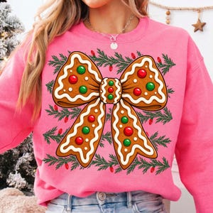 Op de afbeelding: Een felroze sweatshirt met een grote afbeelding van een peperkoekstrik, versierd met wit glazuur en rode en groene snoepjes. Het ontwerp is geplaatst tegen een achtergrond van horizontale groene dennentakken en kleine rode kerstlampjes.