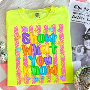 May include: A neon yellow t-shirt with the phrase "Show What You Know" in colorful bubble letters. The shirt features pink striped accents, flowers, stars, and smiley faces, creating a vibrant and playful design.