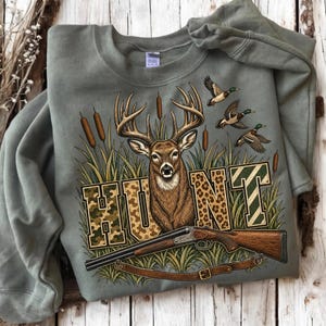 May include: An olive green crewneck sweatshirt featuring a graphic print of a deer head, three flying ducks, and bulrushes. The word HUNT is printed in large block letters with camouflage, leopard, and striped patterns, with a shotgun below.