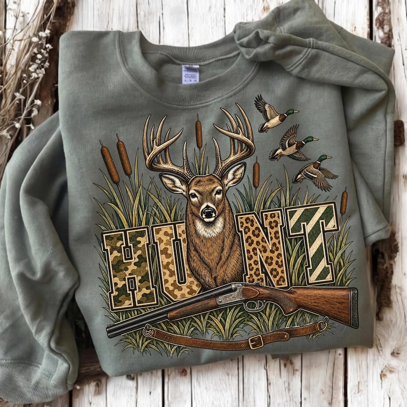 May include: An olive green crewneck sweatshirt featuring a graphic print of a deer head, three flying ducks, and bulrushes. The word HUNT is printed in large block letters with camouflage, leopard, and striped patterns, with a shotgun below.