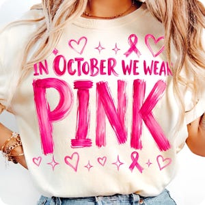 May include: A cream-coloured t-shirt featuring the bright pink text "IN OCTOBER WE WEAR PINK" in a brushstroke font. The design includes pink heart outlines, small four-pointed star icons, and pink awareness ribbons scattered above and below the text.