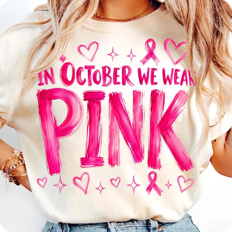 May include: A cream-coloured t-shirt featuring the bright pink text "IN OCTOBER WE WEAR PINK" in a brushstroke font. The design includes pink heart outlines, small four-pointed star icons, and pink awareness ribbons scattered above and below the text.