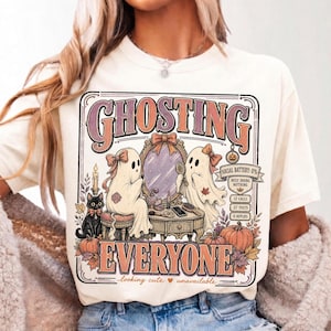 May include: A cream-colored t-shirt featuring a graphic print of two ghosts at a vanity mirror with a black cat, pumpkins, and autumn leaves. The text reads GHOSTING EVERYONE, SOCIAL BATTERY: 0%, BUSY DOING NOTHING, 12 CALLS, 27 TEXTS, 0 REPLIES, looking cute unavailable.
