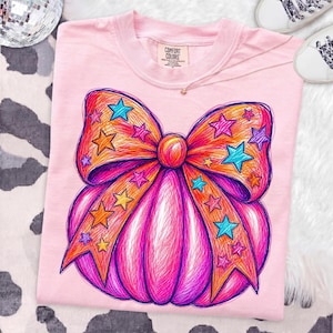 May include: A light pink t-shirt featuring a large, colorful graphic of a pumpkin adorned with a prominent bow. The design is rendered in a scribbled, sketch-like style with vibrant orange, magenta, and purple tones, accented by scattered blue and yellow stars.