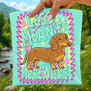 May include: A light mint green fabric pouch featuring a brown dachshund illustration in the center. The text reads LESS MEANIES MORE WEENIES in large light green letters. The design includes pink squiggly borders and small yellow and pink stars.