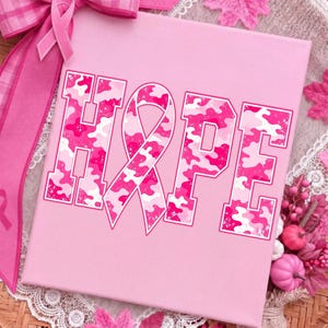 Peut inclure: Une toile carr&eacute;e rose clair affichant le mot HOPE en lettres capitales avec un motif camouflage rose. La lettre O est remplac&eacute;e par un ruban de sensibilisation rose. Le motif est imprim&eacute; dans un style us&eacute; avec des contours blancs sur la surface.