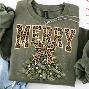 Op de afbeelding: Een olijfgroene sweater met ronde hals en het woord MERRY in grote blokletters met een bruin en zwart luipaardpatroon. Onder de tekst staat een groene dennenboom versierd met een grote luipaardstrik en gouden kerstballen.