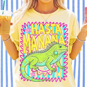 Pu&ograve; includere: Una t-shirt giallo pallido con una stampa grafica di un'iguana verde brillante centrata all'interno di un bordo rosa ondulato. Sopra l'iguana, il testo HASTA MANANA &egrave; stampato in grassetto giallo. Il design include piccole stelle rosa e blu.