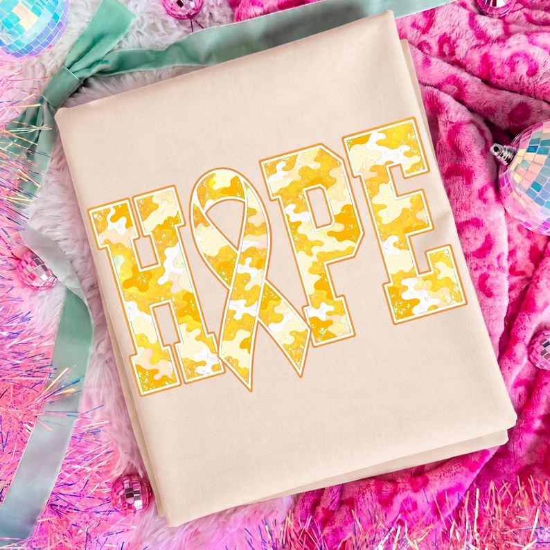 May include: A beige t-shirt featuring the word HOPE printed in a block font. The letter O is replaced by an awareness ribbon. The letters have a yellow and white camouflage pattern with a thin orange outline. The design is centred on the shirt.