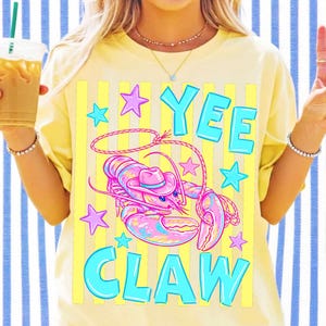 May include: A pale yellow t-shirt featuring a graphic print of a pink lobster wearing a cowboy hat and holding a lasso. The text YEE CLAW is printed in large blue letters above and below the lobster, surrounded by small pink and blue stars.