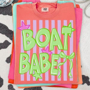 May include: Coral-colored t-shirt with the text "BOAT BABE" in green, accented with stars and a bow. The design is set against a striped background of pink and purple. The shirt is part of a stack of other colorful shirts.