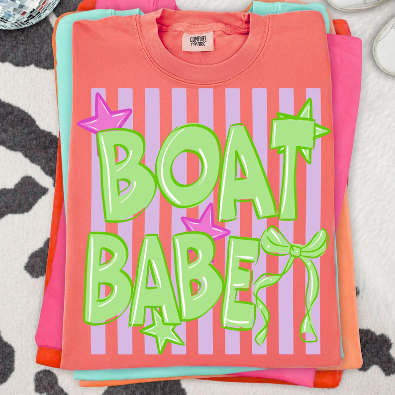 May include: Coral-colored t-shirt with the text "BOAT BABE" in green, accented with stars and a bow. The design is set against a striped background of pink and purple. The shirt is part of a stack of other colorful shirts.