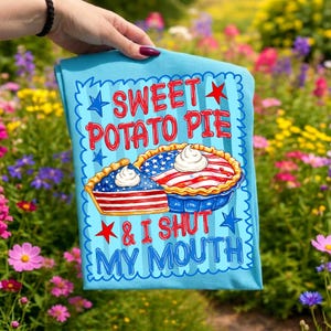 Op de afbeelding: Een lichtblauw t-shirt met een afbeelding van twee taarten met rood, wit en blauwe sterren- en strepenpatronen, getopt met slagroom. De tekst luidt SWEET POTATO PIE & I SHUT MY MOUTH in rode letters, omringd door een blauwe geschulpte rand.