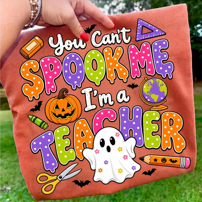 May include: A rust-colored t-shirt featuring the text "You Can't SPOOK ME I'm a TEACHER" in colorful, polka-dotted, dripping letters. The design includes a jack-o'-lantern, a globe, a ghost with flowers, scissors, a pencil, and bats.