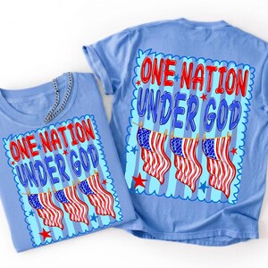 May include: A light blue t-shirt featuring a graphic print with the text ONE NATION UNDER GOD in red and blue letters. The design includes three American flags hanging on a clothesline against a light blue striped background with red and blue stars.