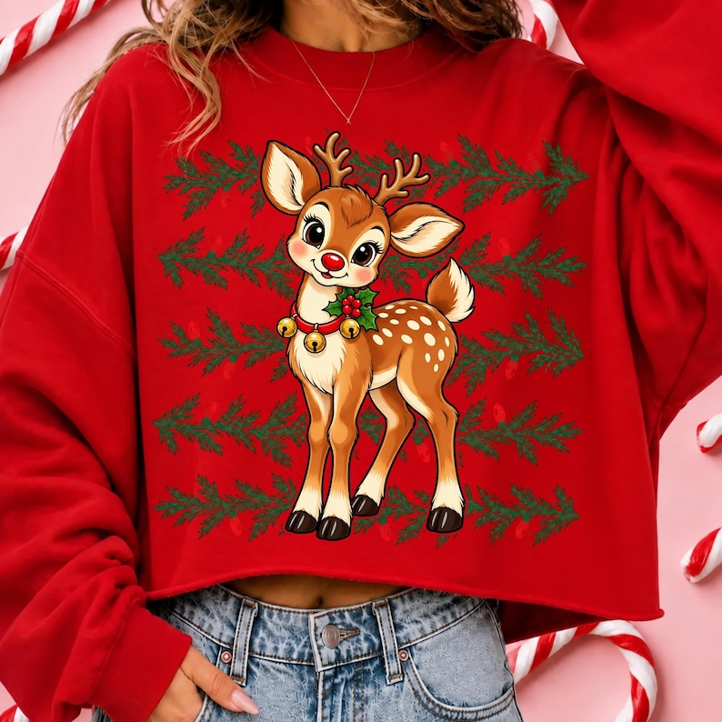 May include: A bright red long-sleeve sweatshirt featuring a central graphic of a brown fawn with a red nose, wearing a collar with gold bells and holly leaves. The background of the graphic displays horizontal rows of green pine branches.