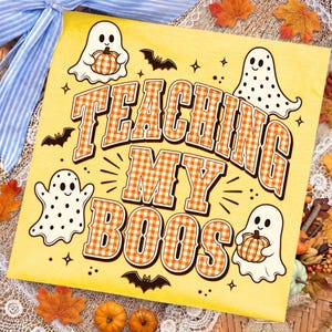 May include: A yellow graphic print featuring the text TEACHING MY BOOS in orange and white gingham letters. The design includes four white ghosts, some with black polka dots or holding orange pumpkins, surrounded by small black bats and stars.