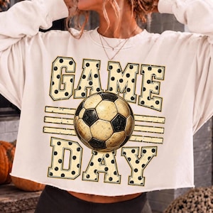 May include: A cream-colored long-sleeve sweatshirt featuring the text "GAME DAY" in large block letters with a black polka dot pattern. A central graphic of a soccer ball with a distressed texture sits between the words "GAME" and "DAY".