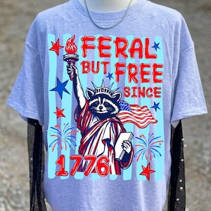 Może przedstawiać: Jasnoszary t-shirt z grafiką szopa pracza przebranego za Statuę Wolności, trzymającego pochodnię i książkę. Napis głosi FERAL BUT FREE SINCE 1776. Wz&oacute;r zawiera czerwone i niebieskie gwiazdy, fajerwerki oraz flagę amerykańską.