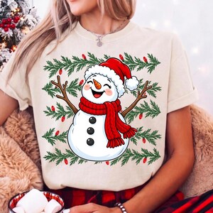 Puede incluir: Una camiseta de color crema con el gr&aacute;fico de un mu&ntilde;eco de nieve sonriente que lleva un gorro y una bufanda rojos. El mu&ntilde;eco de nieve tiene brazos de rama y est&aacute; rodeado por un patr&oacute;n de ramas de pino verdes y peque&ntilde;as luces rojas.