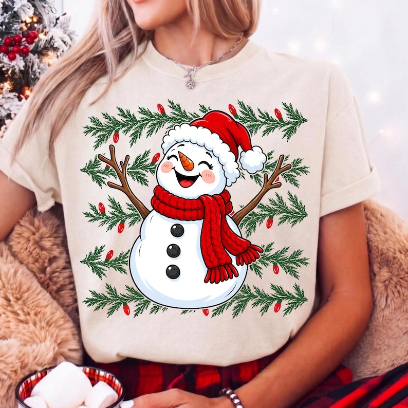 Puede incluir: Una camiseta de color crema con el gr&aacute;fico de un mu&ntilde;eco de nieve sonriente que lleva un gorro y una bufanda rojos. El mu&ntilde;eco de nieve tiene brazos de rama y est&aacute; rodeado por un patr&oacute;n de ramas de pino verdes y peque&ntilde;as luces rojas.