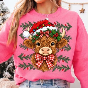Puede incluir: Una sudadera de cuello redondo color rosa brillante con el gr&aacute;fico de una vaca Highland marr&oacute;n que lleva un gorro de Pap&aacute; Noel rojo con pomp&oacute;n blanco, un lazo a cuadros rojos y blancos y una corona de ramas de pino verdes con bayas rojas.