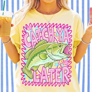 May include: A pale yellow t-shirt featuring a graphic print of a green bass fish centered within a pink, squiggly-bordered frame. The text CATCH YA LATER is printed in large pink letters above and below the fish, accented by small stars.
