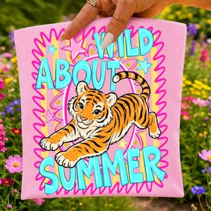 May include: A light pink fabric item featuring a central illustration of a tiger cub. The text reads WILD ABOUT SUMMER in bright blue letters. The design includes pink and yellow striped patterns, star shapes, and a wavy pink border frame.