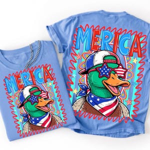 May include: A light blue t-shirt featuring a graphic of a duck wearing a red, white, and blue baseball cap, star-shaped sunglasses, and a matching bandana. The text 'MERICA' is printed in large blue letters above the duck, surrounded by a red squiggly border.