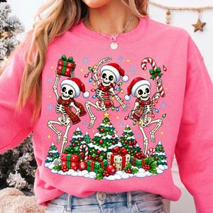 Op de afbeelding: Een felroze sweatshirt met een grafische print van drie dansende skeletten met kerstmutsen en geruite sjaals. De skeletten worden omringd door kerstbomen, ingepakte cadeaus, zuurstokken en kleurrijke kerstverlichting.
