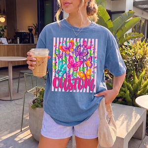 May include: A blue short-sleeve t-shirt featuring a rectangular graphic with vertical white stripes and a cluster of colourful butterflies in shades of pink, orange, yellow, and teal. The word CUSTOM is printed in a bold, cursive pink font below.
