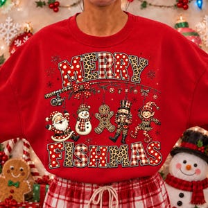 May include: A red crewneck sweatshirt featuring a graphic print with the text MERRY FISHMAS. The design includes a fishing rod hanging ornaments shaped like a Santa, snowman, gingerbread man, nutcracker, and elf, all with leopard and plaid patterns.