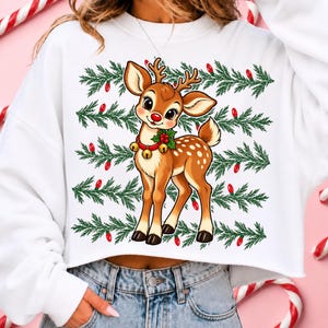 May include: A white cropped sweatshirt featuring a graphic of a brown fawn with a red nose and a collar with gold bells. The background of the graphic consists of horizontal rows of green pine branches interspersed with small red Christmas lights.