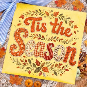 May include: A square yellow sign featuring the text Tis the Season in orange, patterned, and textured lettering. The words are surrounded by a decorative border of embroidered-style autumn leaves, vines, and small floral motifs in shades of orange, red, and green.
