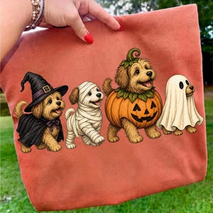 Puede incluir: Un bolso de tela color terracota con un estampado gr&aacute;fico de cuatro perros peque&ntilde;os con disfraces de Halloween. Un perro lleva un sombrero de bruja y una capa negra, otro est&aacute; envuelto en vendas de momia blancas, otro lleva un disfraz de calabaza.