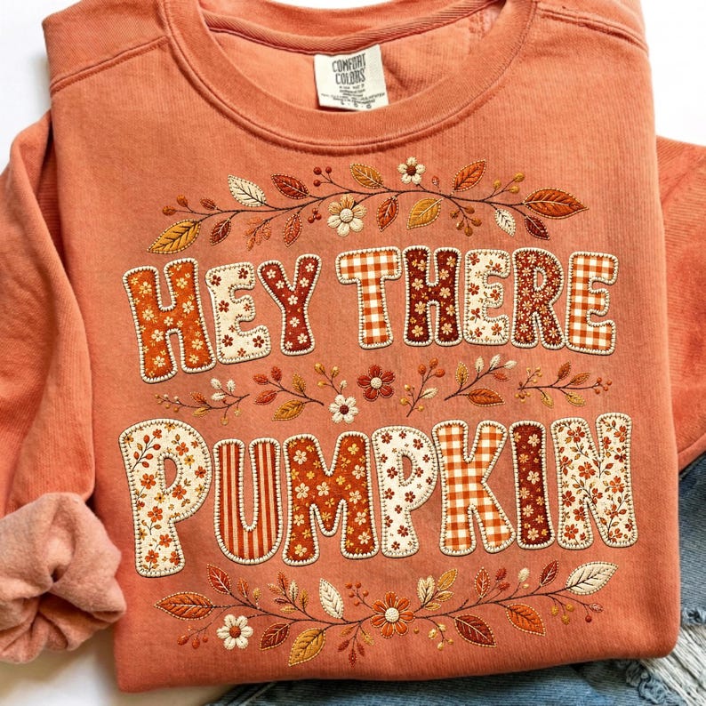 May include: A coral-coloured crewneck sweatshirt featuring the text "HEY THERE PUMPKIN" in large, blocky letters filled with various autumn-themed patterns like orange gingham, floral prints, and stripes, surrounded by embroidered fall foliage.