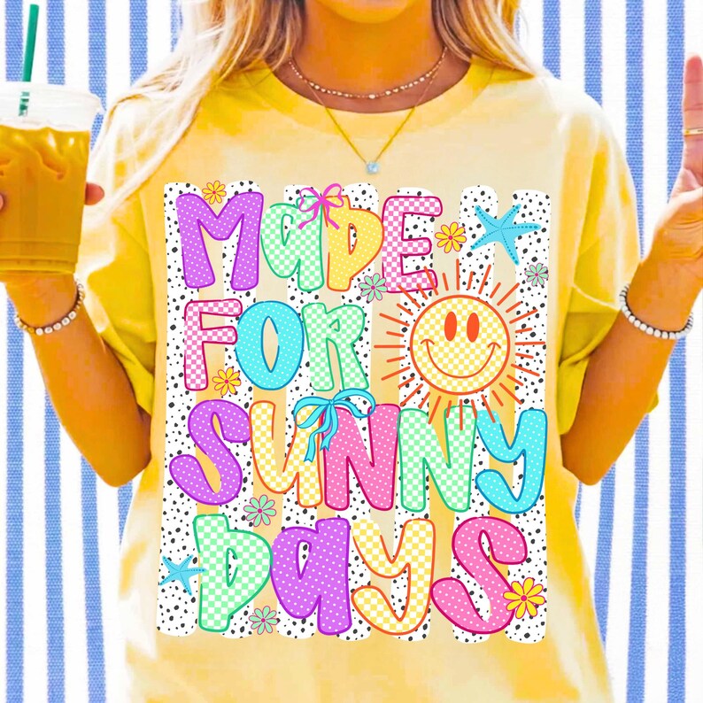 May include: A yellow short-sleeve t-shirt featuring a graphic print with the text "MADE FOR SUNNY DAYS" in colorful, patterned bubble letters. The design includes a yellow smiley face sun, small blue starfish, and flowers on a black dotted background.