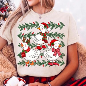 Może przedstawiać: Kremowy t-shirt z grafiką przedstawiającą trzy białe kaczki w świątecznych akcesoriach, takich jak czapki Mikołaja, szaliki i poroże renifera, na tle zielonych gałązek sosnowych z czerwonymi lampkami choinkowymi.